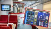 Wnętrze pociągu z automatami, czerwonymi fotelami i z kioskowym barem w tle.