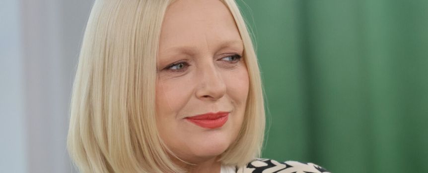 Kobieta z blond włosami i puchem na ramionach, uśmiechająca się na tle zielonej ścianki.