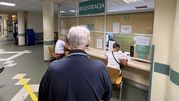 Osoba w ciemnym swetrze czeka w rejestracji w szpitalu, gdzie pracują nieliczni pracownicy.