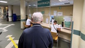 Osoba w ciemnym swetrze czeka w rejestracji w szpitalu, gdzie pracują nieliczni pracownicy.