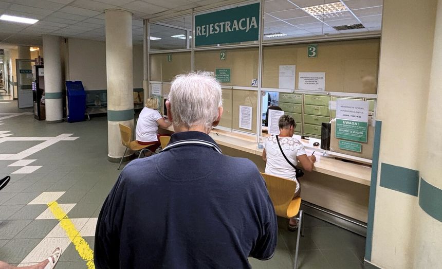 Osoba w ciemnym swetrze czeka w rejestracji w szpitalu, gdzie pracują nieliczni pracownicy.