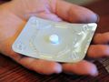 Na zdjęciu dłoń trzyma opakowanie tabletki, na które nałożona jest biały tablet.