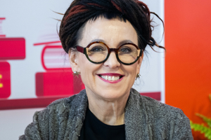 Na zdjęciu uśmiechnięta kobieta z okularami i długimi włosami, w szarym swetrze, na tle książek.