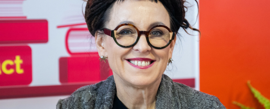 Na zdjęciu uśmiechnięta kobieta z okularami i długimi włosami, w szarym swetrze, na tle książek.