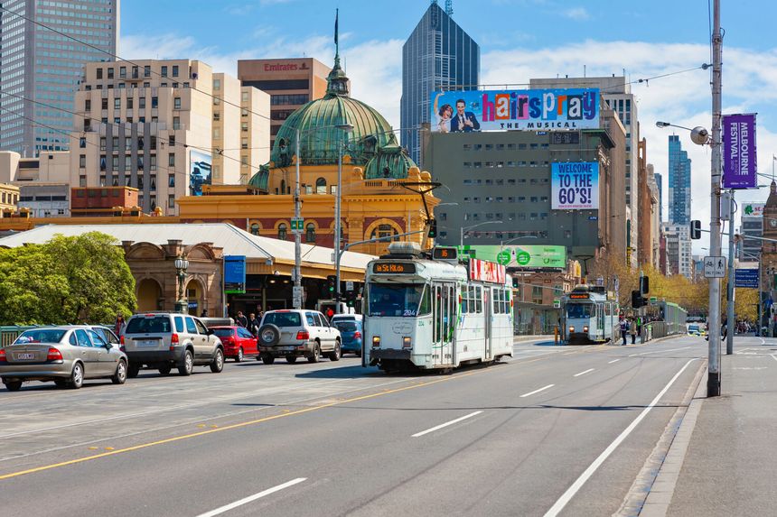 Na zdjęciu ulica w mieście, tramwaje, budynki i reklamy. Dzień słoneczny, ruch uliczny.
