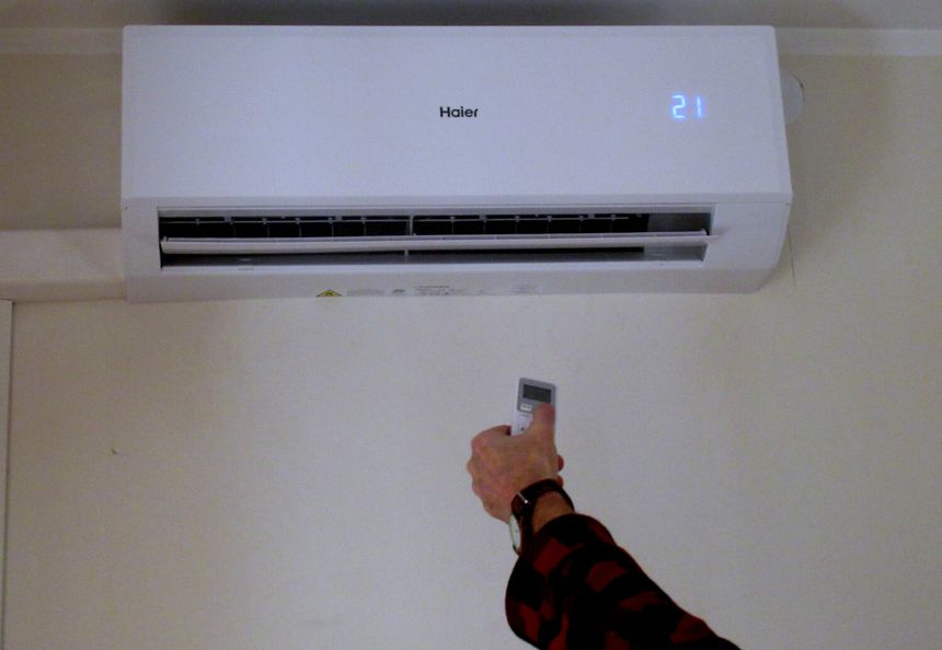 Klimatyzator na ścianie, ręka trzymająca pilot z ustawioną temperaturą 21 stopni.