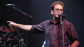 Mężczyzna z mikrofonem śpiewa na scenie, w tle instrumenty muzyczne i światła.