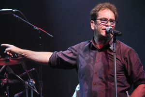 Mężczyzna z mikrofonem śpiewa na scenie, w tle instrumenty muzyczne i światła.