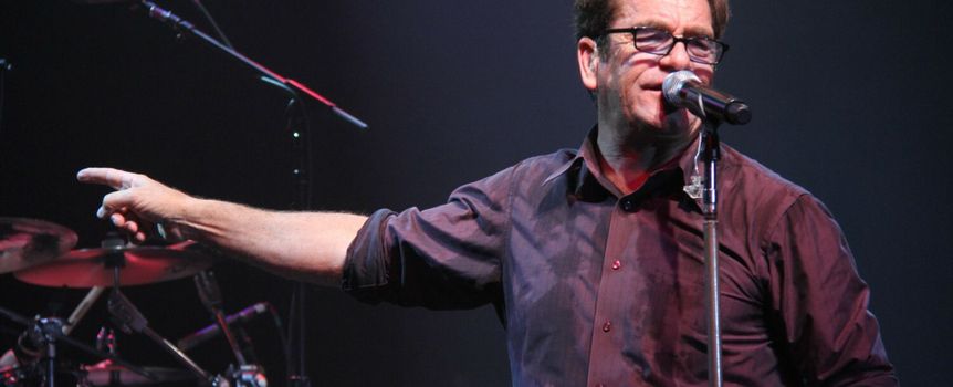 Mężczyzna z mikrofonem śpiewa na scenie, w tle instrumenty muzyczne i światła.