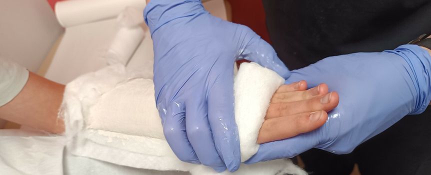 Zdjęcie przedstawia rękę opatrywaną bandażem przez osobę w niebieskich rękawiczkach.
