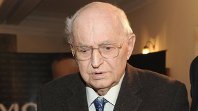 Osoba starsza o okularach, w ciemnym garniturze, zmartwiona w towarzystwie innych.