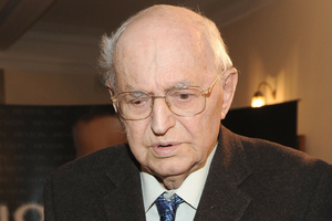 Osoba starsza o okularach, w ciemnym garniturze, zmartwiona w towarzystwie innych.