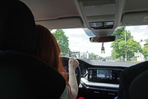 Kobieta prowadzi samochód, widok z tylnego siedzenia na drogę i drzewa po bokach.