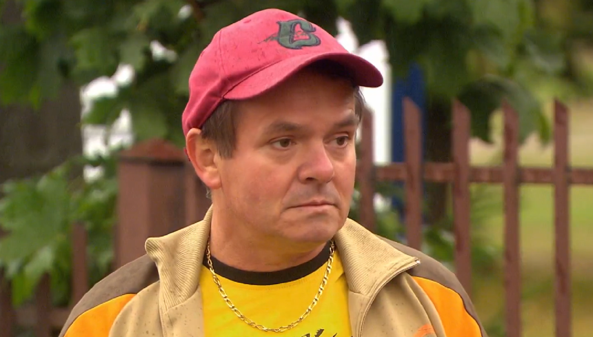 Mężczyzna w czerwonej czapce, ubrany w żółtą koszulę i beżową kurtkę, stoi na tle zieleni.