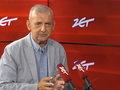 Mężczyzna w jasnym garniturze, mówi do mikrofonów na tle czerwonej ściany z logo.