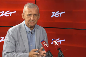 Mężczyzna w jasnym garniturze, mówi do mikrofonów na tle czerwonej ściany z logo.