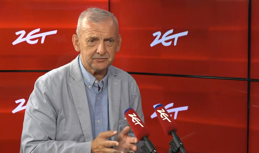 Mężczyzna w jasnym garniturze, mówi do mikrofonów na tle czerwonej ściany z logo.