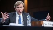 Mężczyzna w garniturze mówi do mikrofonu, gestykulując rękami. Na stole butelka wody.