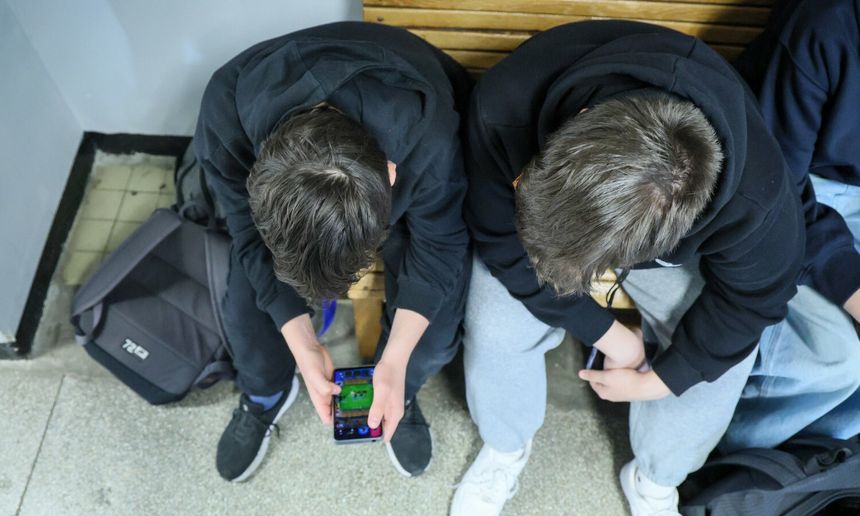 Dwa chłopcy w czarnych bluzach siedzą na ławce, jeden trzyma telefon, grając w grę.
