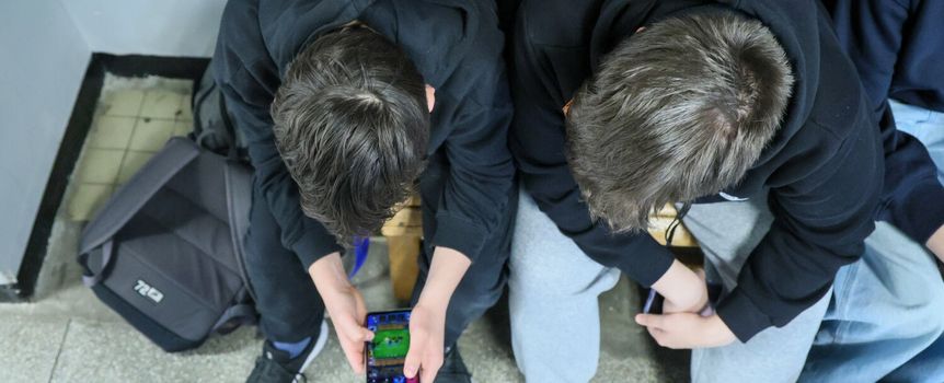 Dwa chłopcy w czarnych bluzach siedzą na ławce, jeden trzyma telefon, grając w grę.