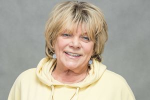 Kobieta w żółtej bluzie z kapturem, uśmiechnięta, z krótkimi włosami. Tło szare.