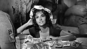 Gina Lollobrigida. Kobieta w ciemnej bluzce, otoczona banknotami, patrzy z uśmiechem w stronę obiektywu.