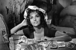 Gina Lollobrigida. Kobieta w ciemnej bluzce, otoczona banknotami, patrzy z uśmiechem w stronę obiektywu.