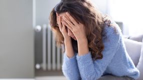 Kobieta z długimi, kręconymi włosami, w niebieskim swetrze, zasłania twarz dłońmi, wygląda na zmartwioną.