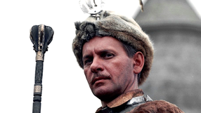 Mężczyzna w futrzanej czapce, trzymający berło, w tle zamek. Historyczny lub filmowy motyw.