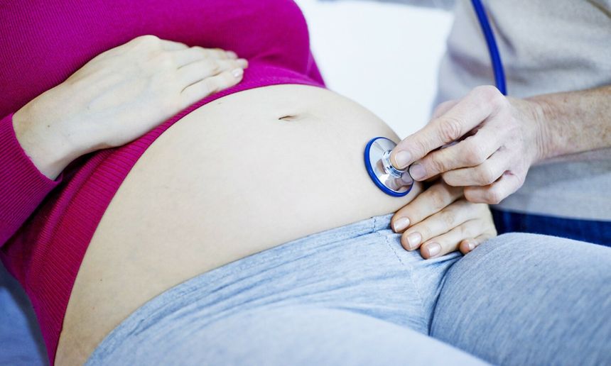 Kobieta w ciąży trzyma rękę na brzuchu, lekarz słucha stethoskopem.
