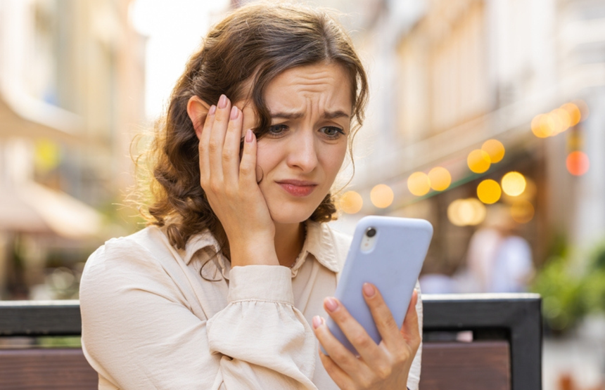 Kobieta zmartwiona, trzymająca telefon, siedzi na ławce w mieście. W tle mgliste światła.