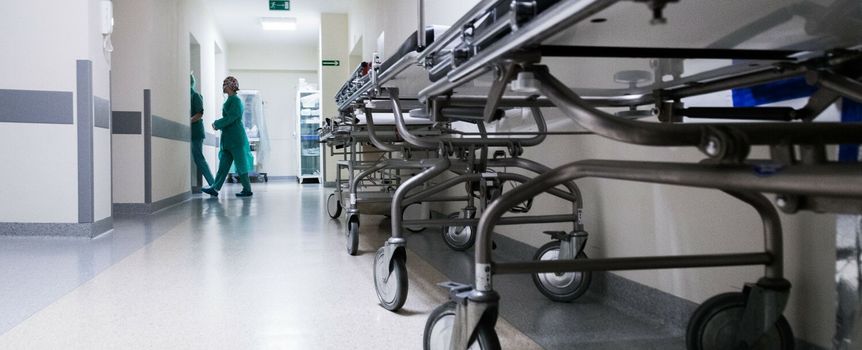 Korytarz szpitalny z metalowymi łóżkami i pielęgniarką w zielonym stroju.