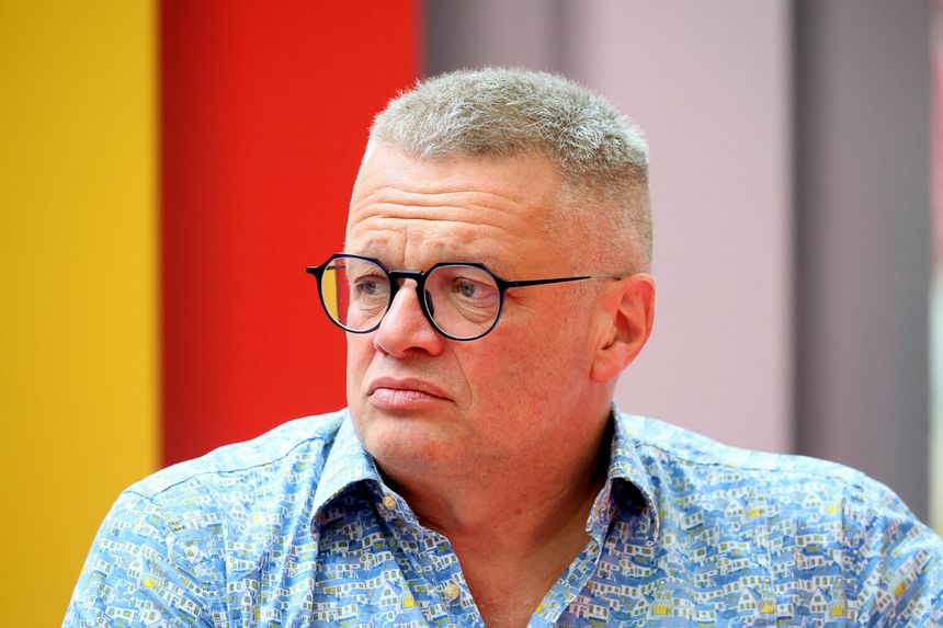 Marcin Meller z krótkimi włosami i okrągłymi okularami, ubrany w kolorową koszulę. Tło w żywych kolorach.