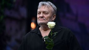 Mężczyzna z siwym włosami, w czarnej koszuli, trzyma białą różę. Zdjęcie w plenerze, lekko rozmazane tło.