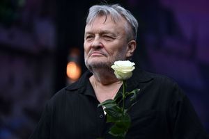 Mężczyzna z siwym włosami, w czarnej koszuli, trzyma białą różę. Zdjęcie w plenerze, lekko rozmazane tło.