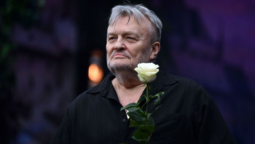 Mężczyzna z siwym włosami, w czarnej koszuli, trzyma białą różę. Zdjęcie w plenerze, lekko rozmazane tło.