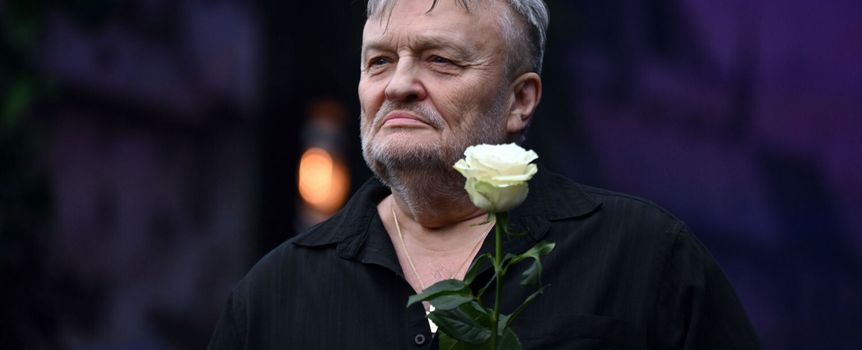 Mężczyzna z siwym włosami, w czarnej koszuli, trzyma białą różę. Zdjęcie w plenerze, lekko rozmazane tło.