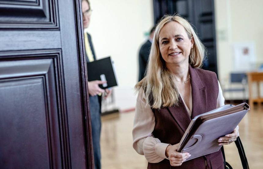 Na zdjęciu uśmiechająca się kobieta w brązowej kamizelce trzyma teczkę. W tle mężczyzna w garniturze.