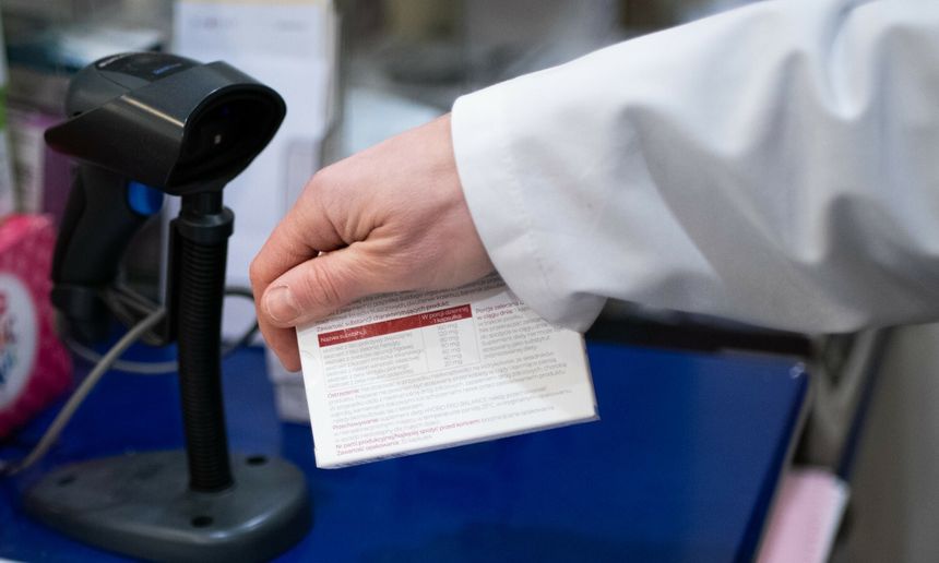 Osoba w białym fartuchu trzyma kartonik z informacjami. W tle skaner kodów kreskowych.