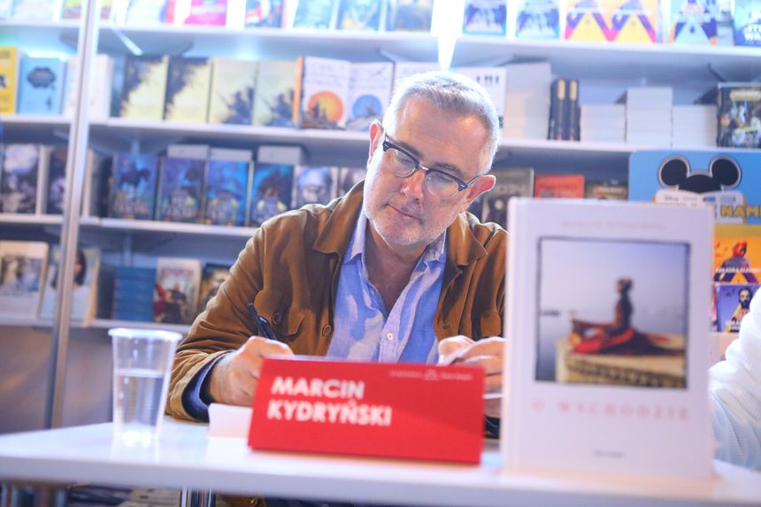 Mężczyzna przy stoliku podpisuje książki. W tle regał z książkami. To Marcin Kydryński.