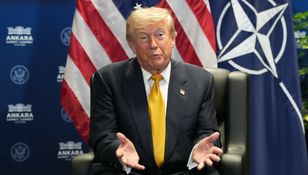 Na zdjęciu mężczyzna w garniturze z żółtym krawatem, siedzący na tle flag USA i NATO.