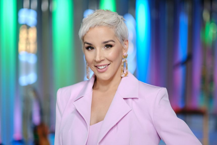 Uśmiechnięta Lil Masti w różowej marynarce na tle kolorowych świateł, z długimi kolczykami.