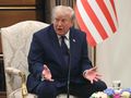 Mężczyzna w garniturze siedzi na krześle, gestykulując podczas rozmowy. W tle flaga USA.