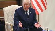 Mężczyzna w garniturze siedzi na krześle, gestykulując podczas rozmowy. W tle flaga USA.