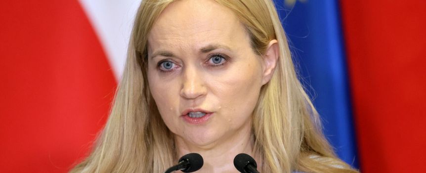 Kobieta z blond włosami przemawia do mikrofonu, w tle flagi Polski i UE.