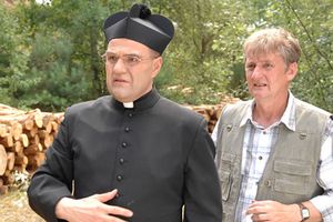 Mężczyźni w strojach zwyczajnych i duchownym, w tle stosy drewna i zieleń lasu.