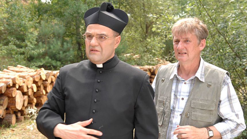 Mężczyźni w strojach zwyczajnych i duchownym, w tle stosy drewna i zieleń lasu.