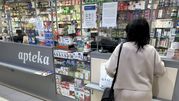 Kobieta w jasnym stroju stoi przy ladzie apteki, otoczonej lekami i suplementami.