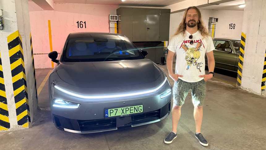 Mężczyzna stoi obok nowoczesnego samochodu w garażu. Na sobie ma koszulkę z logo zespołu Metallica.