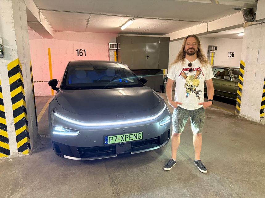 Mężczyzna w t-shircie Metallica stoi obok nowoczesnego samochodu w parkingu.